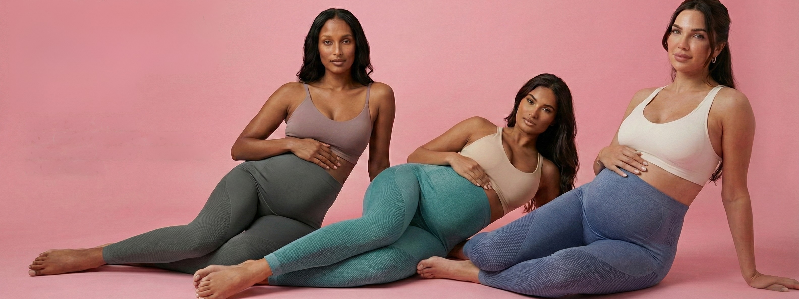 Three women sitting on a pink background wearing colorful leggings and sports bras.